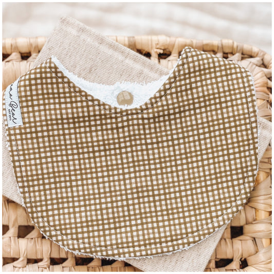 Forest green gingham baby bib with a white grid pattern, styled on a woven basket with a neutral linen cloth.