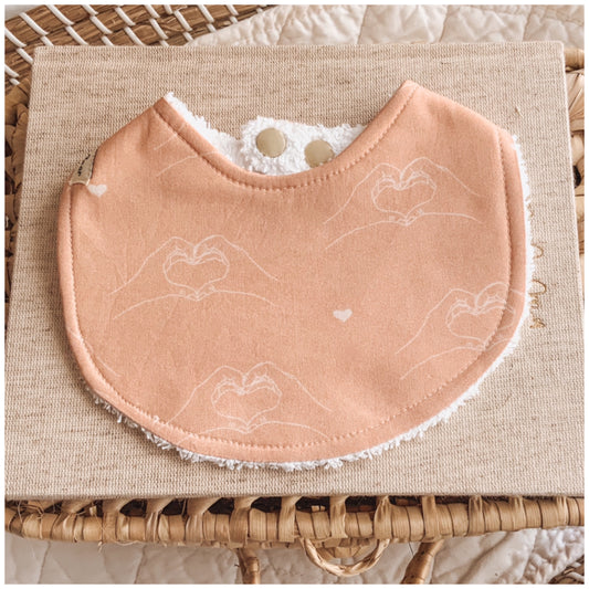 Handmade baby bib in terracotta with delicate line drawings of hands forming heart shapes, backed with white terry towelling, displayed on neutral fabric with a natural woven basket background