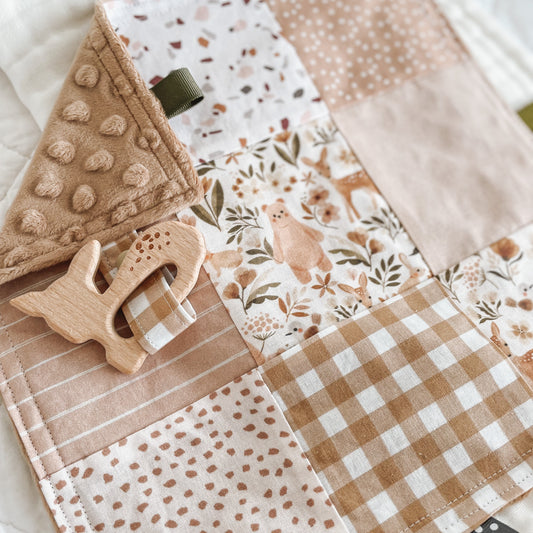 Handmade baby Taggie blanket in warm earthy tones with a mix of patchwork prints including gingham, florals, terrazzo, stripes, and woodland animals. Features a wooden deer-shaped teether attached with a matching fabric strap, textured minkee backing, and neutral-toned ribbon loops for sensory play.