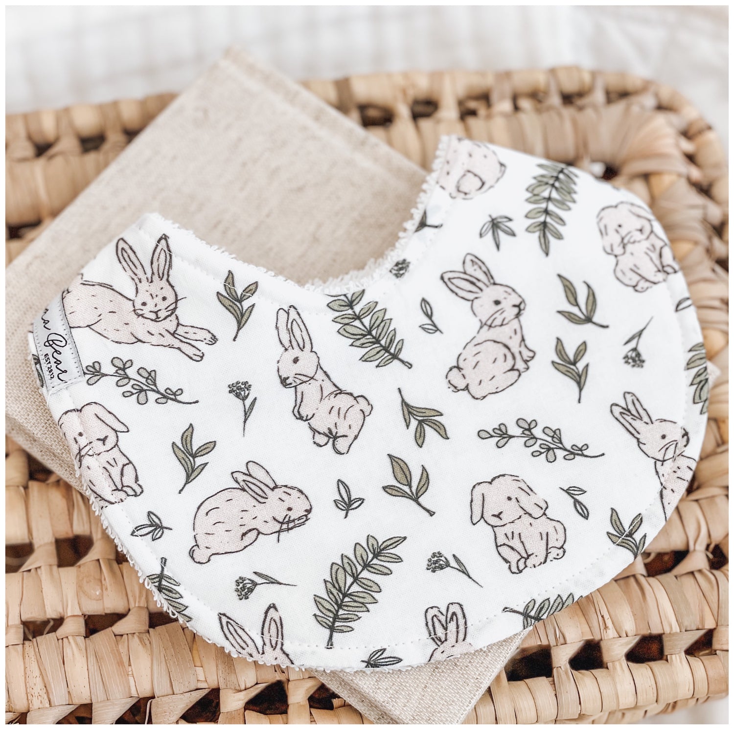 Handmade baby bib featuring light brown bunny illustrations and green leafy branches on a white background. Bib is backed with beige terry towelling and displayed on a woven basket.