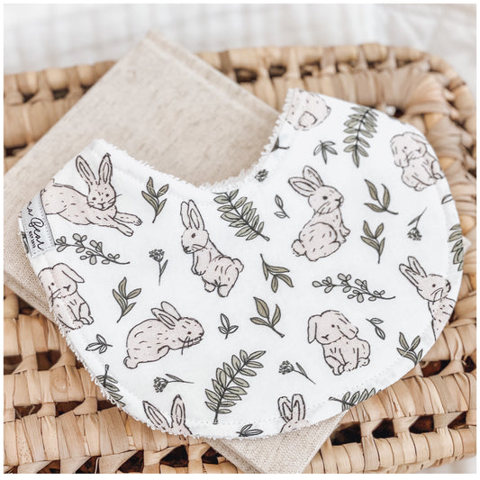 Handmade baby bib featuring light brown bunny illustrations and green leafy branches on a white background. Bib is backed with beige terry towelling and displayed on a woven basket.