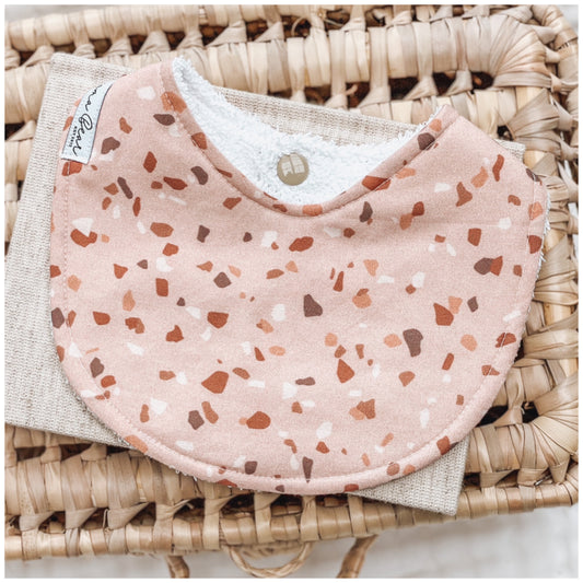 Peach baby bib with scattered terrazzo-style spots in warm neutral tones, displayed on beige linen and a woven basket.