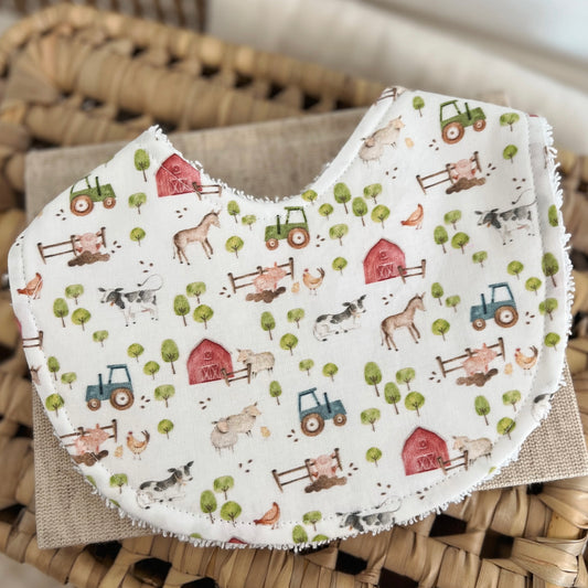 Flatlay of a handmade baby bib with a farm-themed pattern. The design includes cows, sheep, horse, chickens, blue and green tractors, red barns, and small trees on a light cream base. The bib has a cotton front and a soft terry towelling back for absorbency.