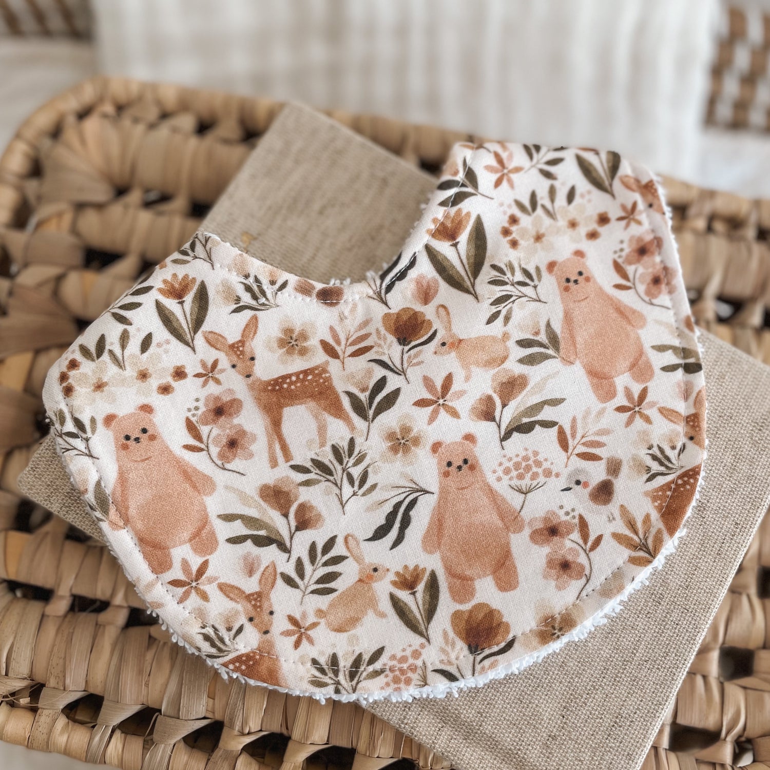 Neutral baby bib with a woodland animal pattern including bears, deer, bunnies, and florals in warm tan, brown, and rust tones on a light background. Absorbent terry backing ideal for newborn messes and teething.