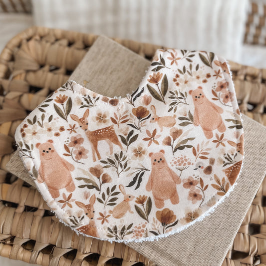 Neutral baby bib with a woodland animal pattern including bears, deer, bunnies, and florals in warm tan, brown, and rust tones on a light background. Absorbent terry backing ideal for newborn messes and teething.