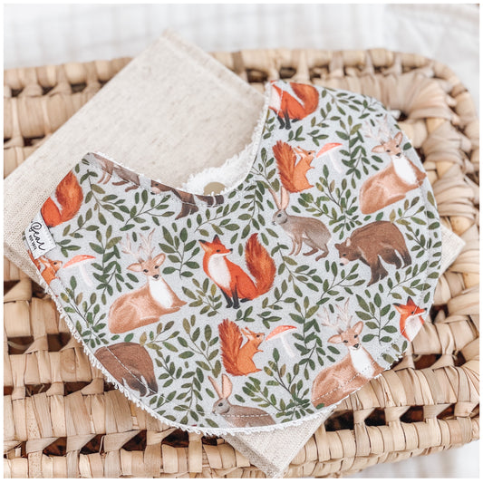 Handmade baby bib with a woodland forest print showing foxes, deer, bears, squirrels, and rabbits surrounded by green leaves on a light background. Bib is backed with beige terry towelling and displayed on a woven basket.