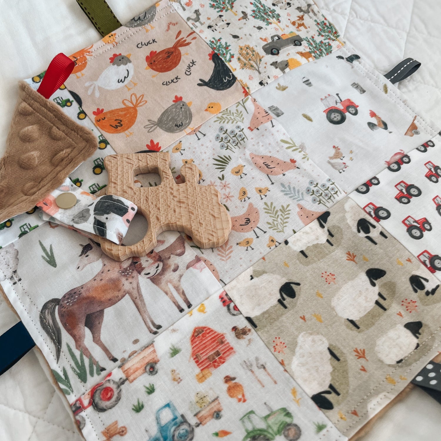 Handmade baby Taggie blanket with a farmyard patchwork design featuring horses, sheep, chickens, tractors, barns, and farm animals in earthy tones. Includes a wooden cow-shaped teether attached with a cow print fabric strap, minkee dot backing, and a mix of colourful ribbon loops.
