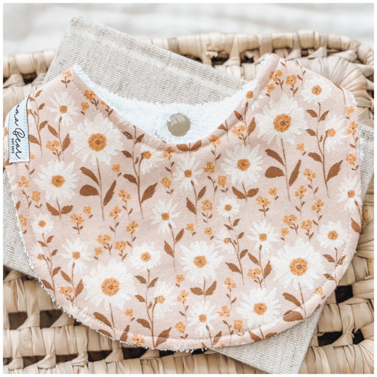 Peach-toned floral baby bib with white daisies and golden sprigs, displayed on a woven basket background with a neutral cloth underneath.