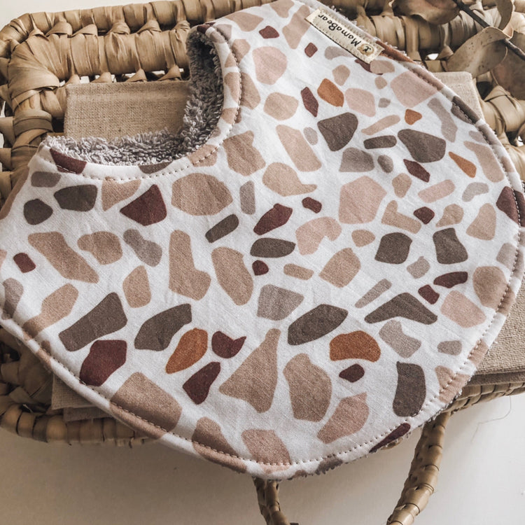Modern baby bib with terrazzo-style print in warm earthy tones and soft white terry towelling backing.