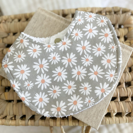Flatlay of a handmade baby bib with a soft green-grey background and white daisy print. The daisies have peachy orange centres and are evenly spaced across the fabric. Bib is made from cotton and backed with absorbent terry towelling.