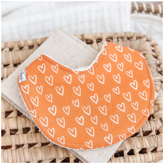 Handmade baby bib with a bright orange background and scattered white heart pattern. Bib is backed with beige terry towelling and displayed on a woven basket.