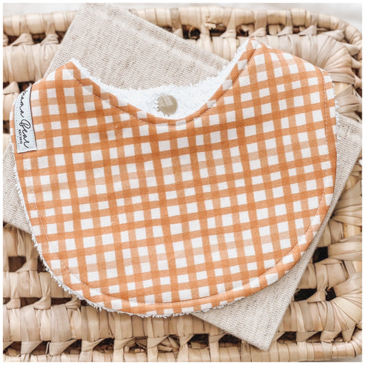 Terracotta gingham check baby bib with soft white squares, displayed on a natural woven basket and neutral cloth.