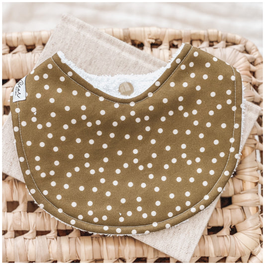 Sage green baby bib with a white polka dot print, styled on a woven basket with a neutral cloth underneath.
