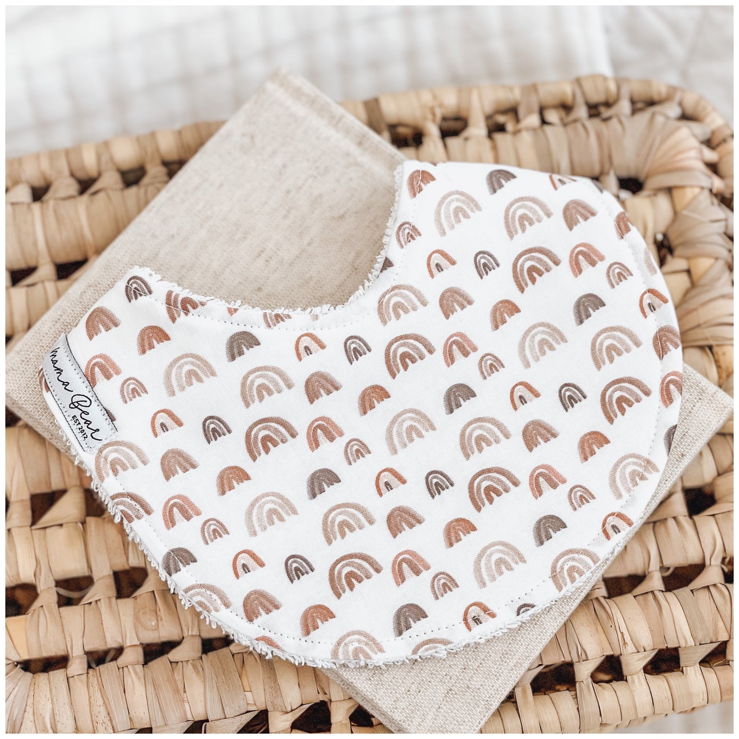 Neutral baby bib with warm-toned rainbow print in rust, beige, and brown shades, styled on a linen cloth over a woven basket.