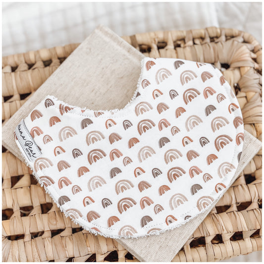 Neutral baby bib with warm-toned rainbow print in rust, beige, and brown shades, styled on a linen cloth over a woven basket.