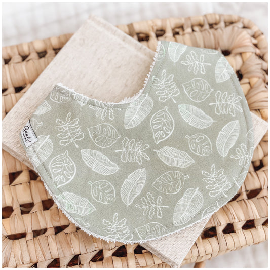 Handmade baby bib with a sage green background and white outlined botanical leaf illustrations. Bib is backed with beige terry towelling and styled on a woven basket.