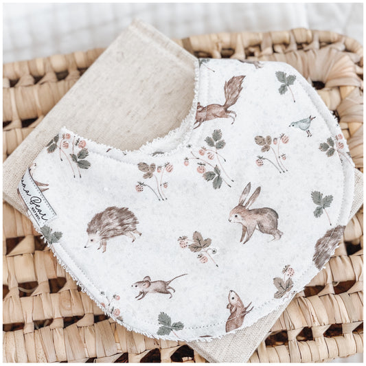  Handmade baby bib with an illustrated woodland animal print, including rabbits, hedgehogs, mice, squirrels, birds, and strawberry vines on a soft cream background. Bib is laid on a beige cloth over a woven basket.