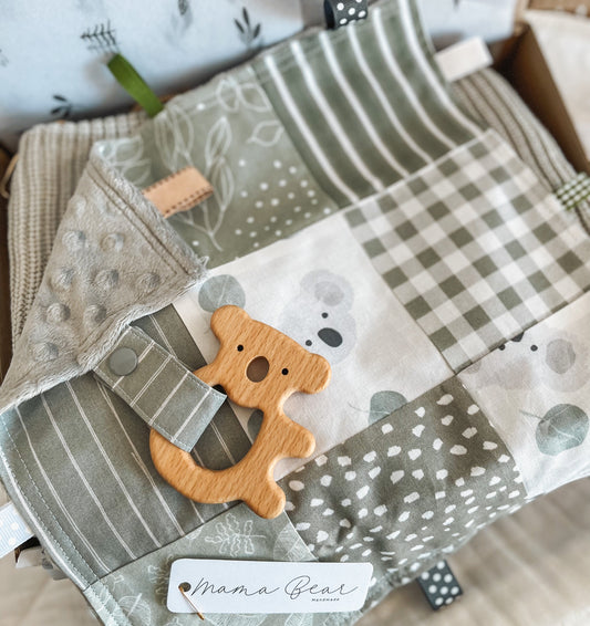 Handmade baby Taggie blankie in sage green tones, featuring a mix of gingham, stripes, polka dots, and soft koala print fabric. Includes a wooden koala teether attached with a matching fabric strap, textured backing, and multiple ribbon loops. Finished with a “Mama Bear Handmade” tag.