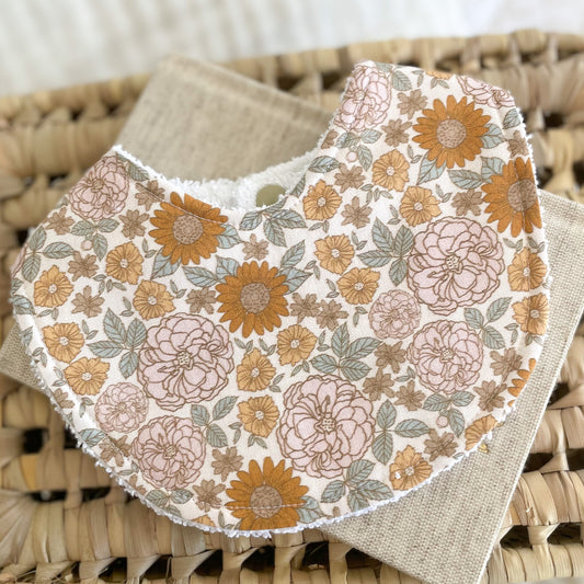 Retro floral baby bib with mustard, blush, and beige flowers on an off-white background. Bib is laid flat on a woven mat with a neutral linen cloth underneath.